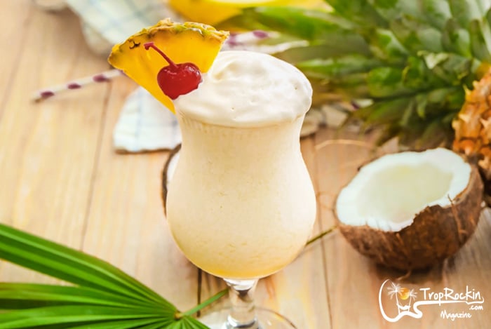 A piña colada garnished with a pineapple slice and a cherry in a curved glass, placed on a wooden surface with a coconut and pineapple in the background.