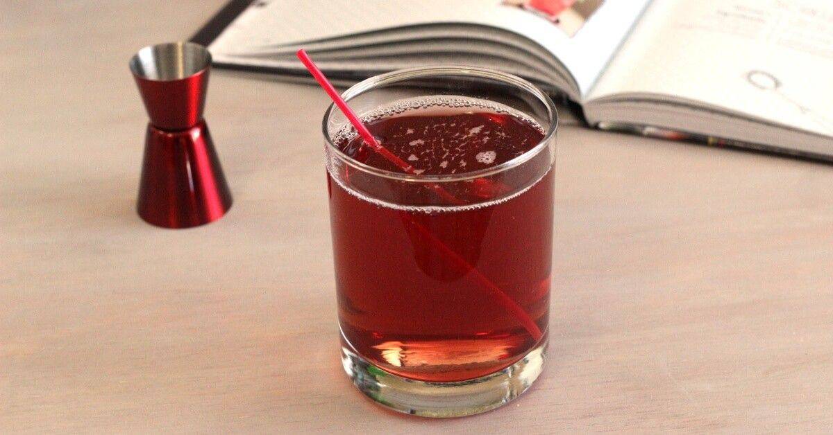 A glass of reddish-brown beverage with a red straw, sitting on a light table next to a red jigger and an open book.