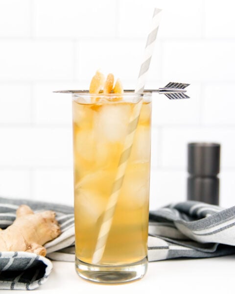 A tall glass filled with iced amber-colored liquid, garnished with candied ginger, a striped straw, and an arrow pick, sits on a striped cloth with ginger root nearby.