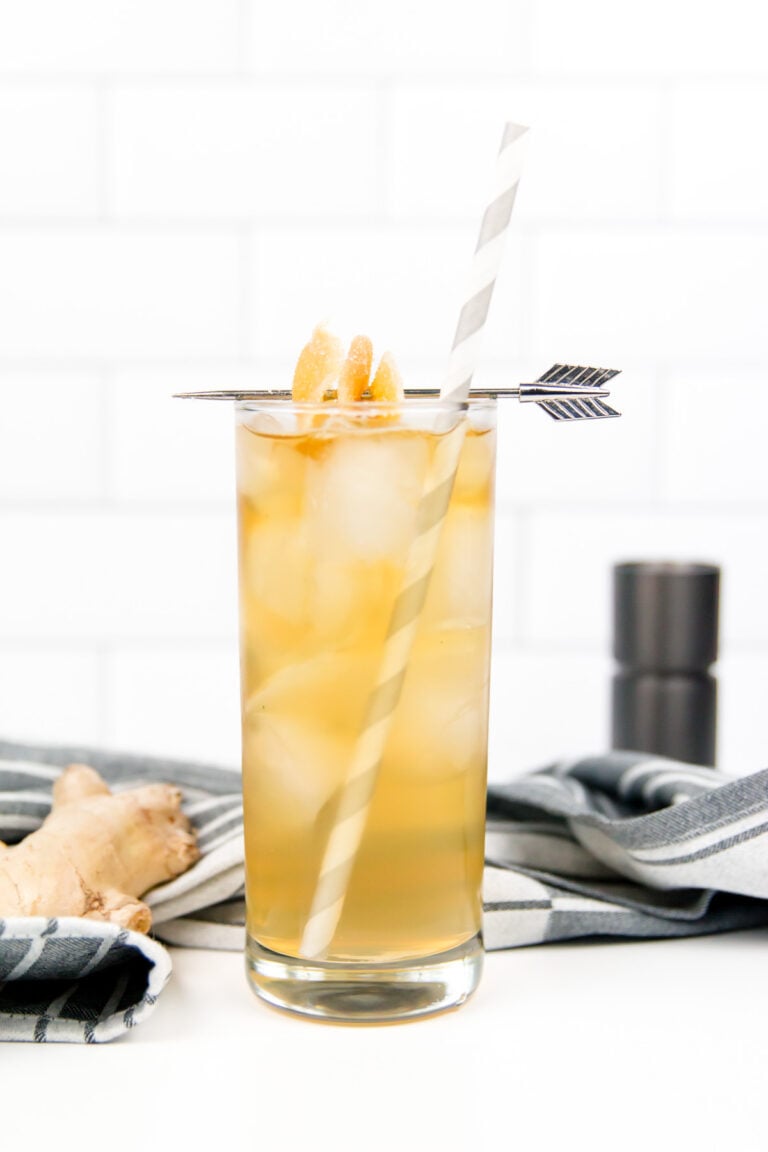 A tall glass filled with iced amber-colored liquid, garnished with candied ginger, a striped straw, and an arrow pick, sits on a striped cloth with ginger root nearby.
