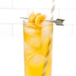 A tall glass filled with iced orange beverage, garnished with sugared orange peel and a striped straw, against a white tiled background.