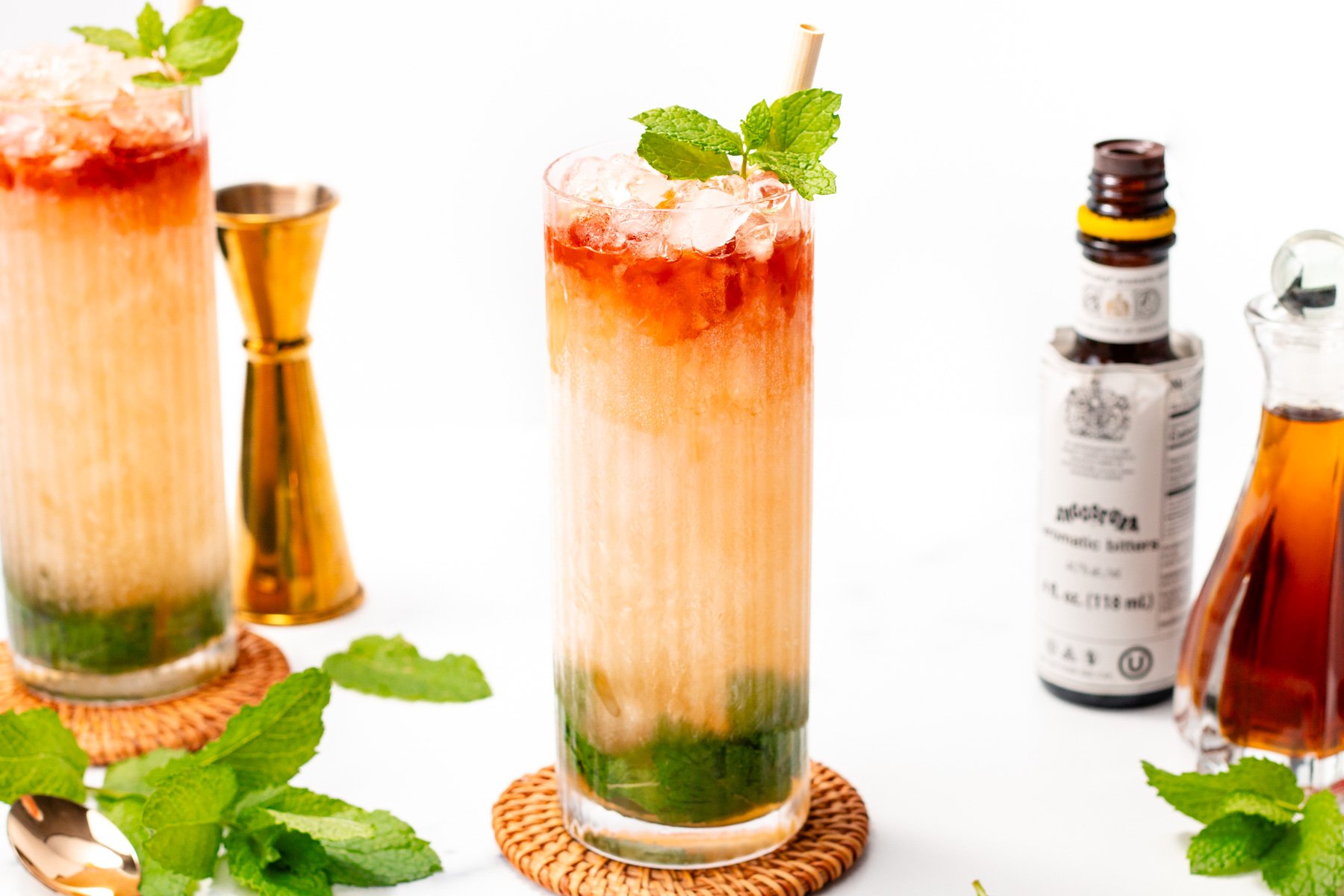 A glass of mint julep cocktail with crushed ice, mint leaves, and a straw, accompanied by bitters, a jigger, and a small carafe on a white surface.