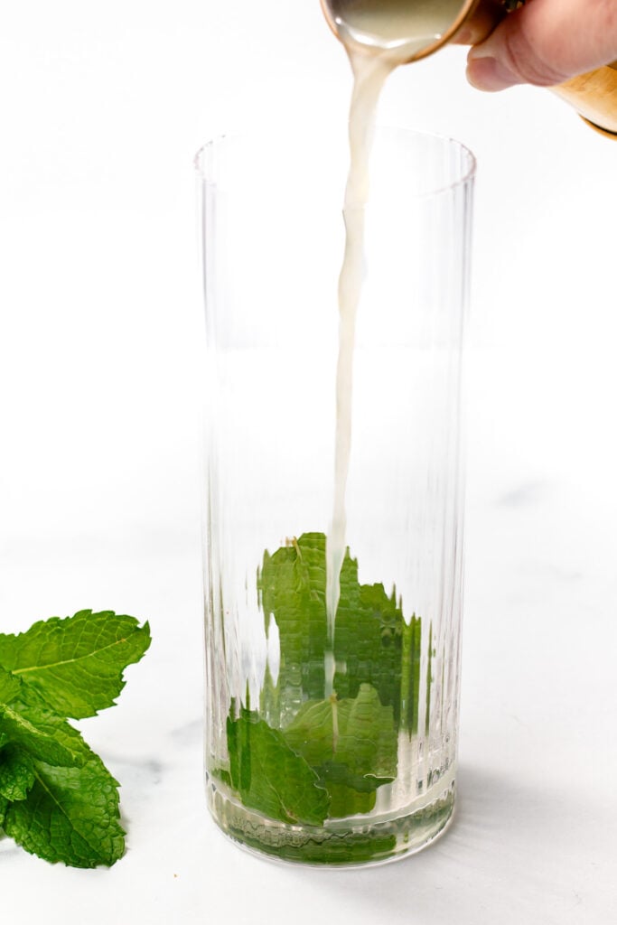 A hand pours liquid into a tall glass containing fresh mint leaves, with additional mint leaves placed on the white surface nearby.