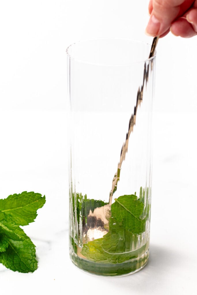 A hand uses a muddler in a tall glass with fresh mint leaves at the bottom; extra mint leaves lie on the surface beside the glass.