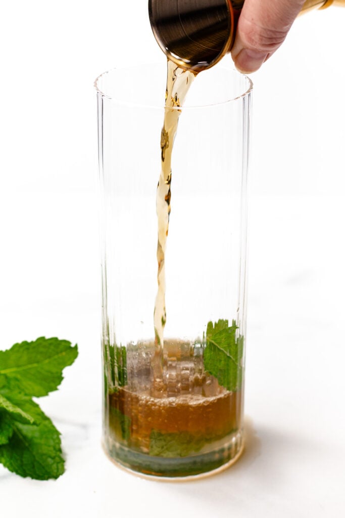 A liquid is being poured from a metal jigger into a tall glass containing mint leaves and a brown mixture, with a mint sprig placed beside the glass.