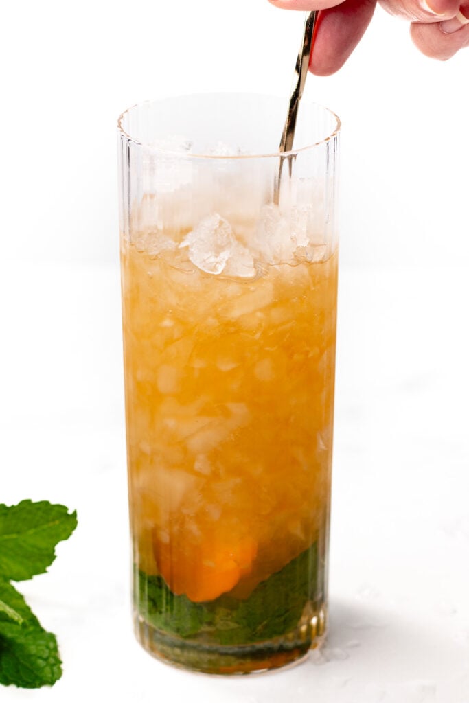 A hand stirs a tall glass filled with crushed ice, brown liquid, and mint leaves at the bottom.