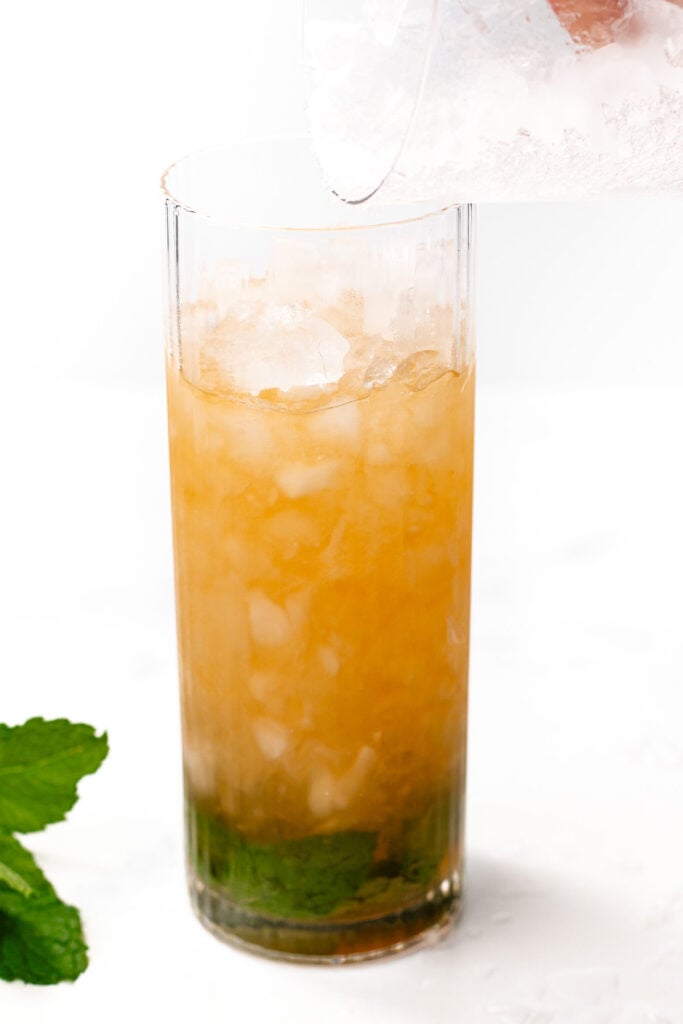 A tall glass with crushed ice, brown liquid, and muddled green mint leaves at the bottom. A hand pours more crushed ice into the glass. Mint leaves are nearby.