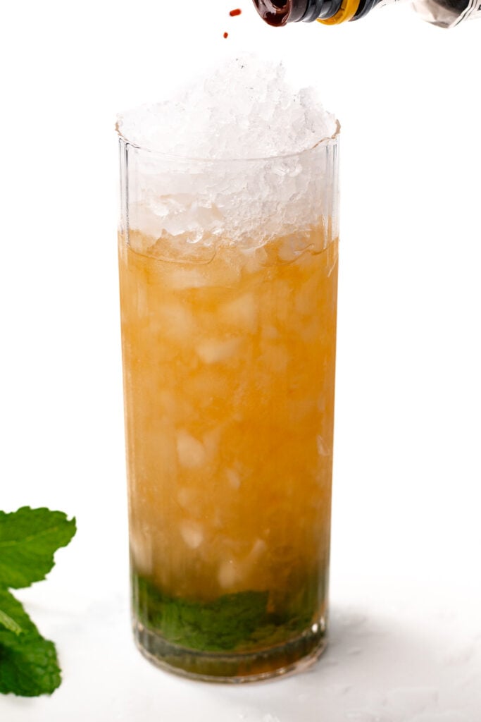 A tall glass with crushed ice, amber-colored liquid, and mint leaves at the bottom. A dark liquid is being poured from a bottle onto the ice. Mint leaves are beside the glass.