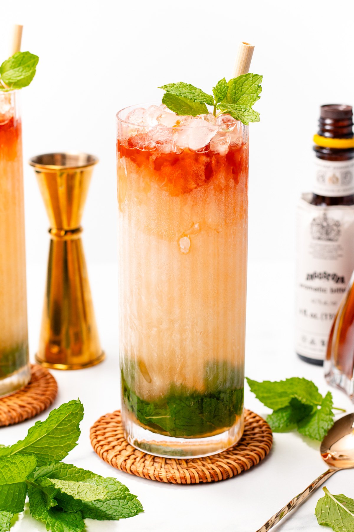 A tall glass filled with a layered cocktail, crushed ice, fresh mint, and a straw, set on a woven coaster with bar tools and bitters in the background.