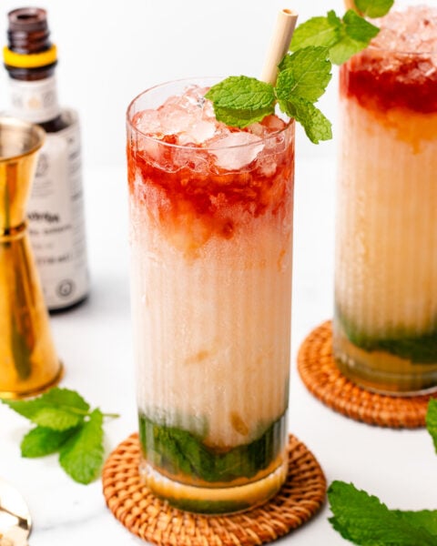 A tall glass filled with a layered iced drink featuring green, creamy, and red sections, garnished with fresh mint and a straw, sitting on a woven coaster.