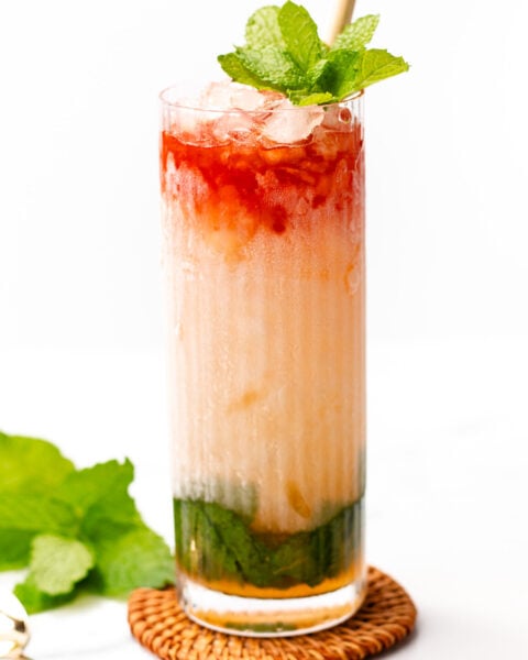 A tall glass of colorful layered iced drink with mint leaves on top, a straw, and crushed ice, placed on a wicker coaster, mint leaves beside the glass.