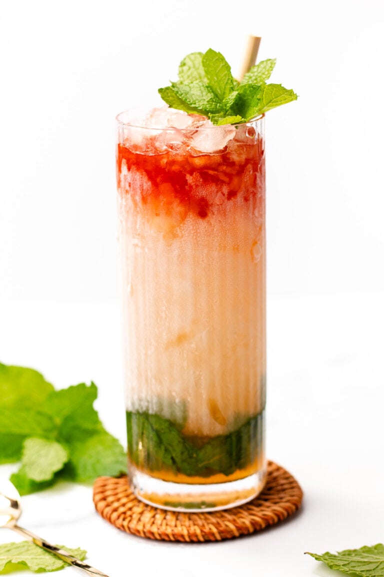 A tall glass of colorful layered iced drink with mint leaves on top, a straw, and crushed ice, placed on a wicker coaster, mint leaves beside the glass.