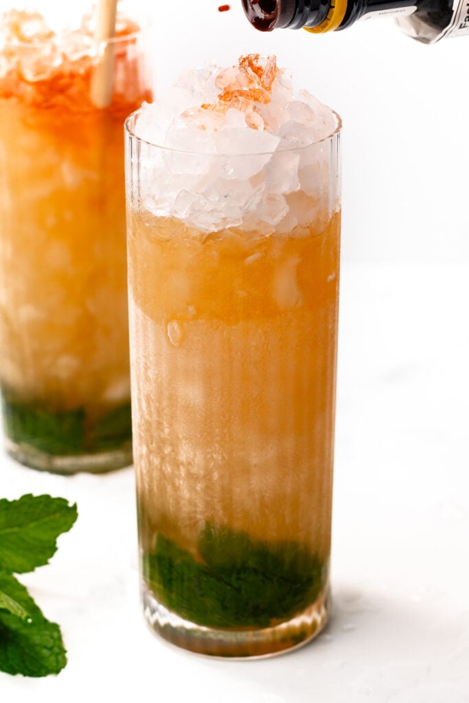 A highball glass filled with crushed ice, mint leaves at the bottom, and an amber cocktail being topped with bitters. Another similar drink and mint garnish are in the background.