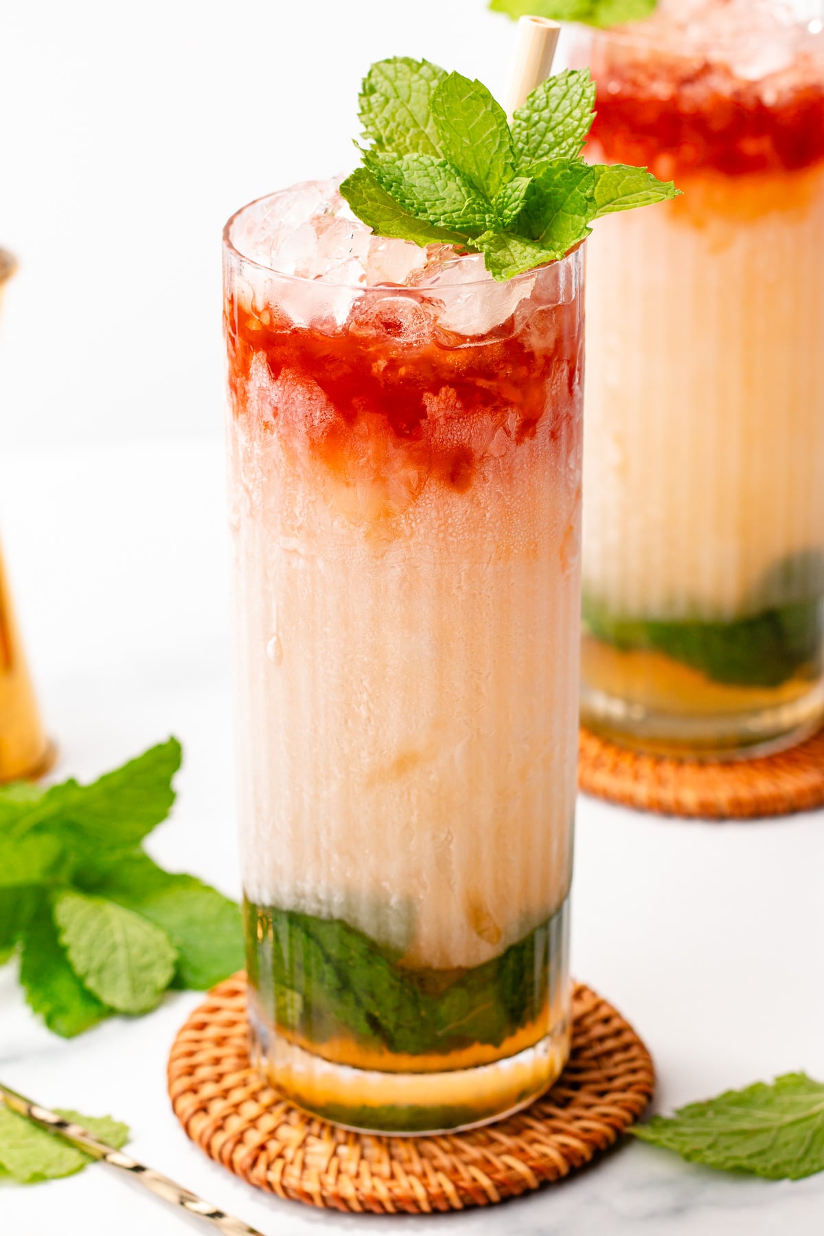 A tall glass of iced layered drink with mint leaves, green syrup at the bottom, creamy middle layer, and a red top layer, garnished with fresh mint and a straw, on a woven coaster.