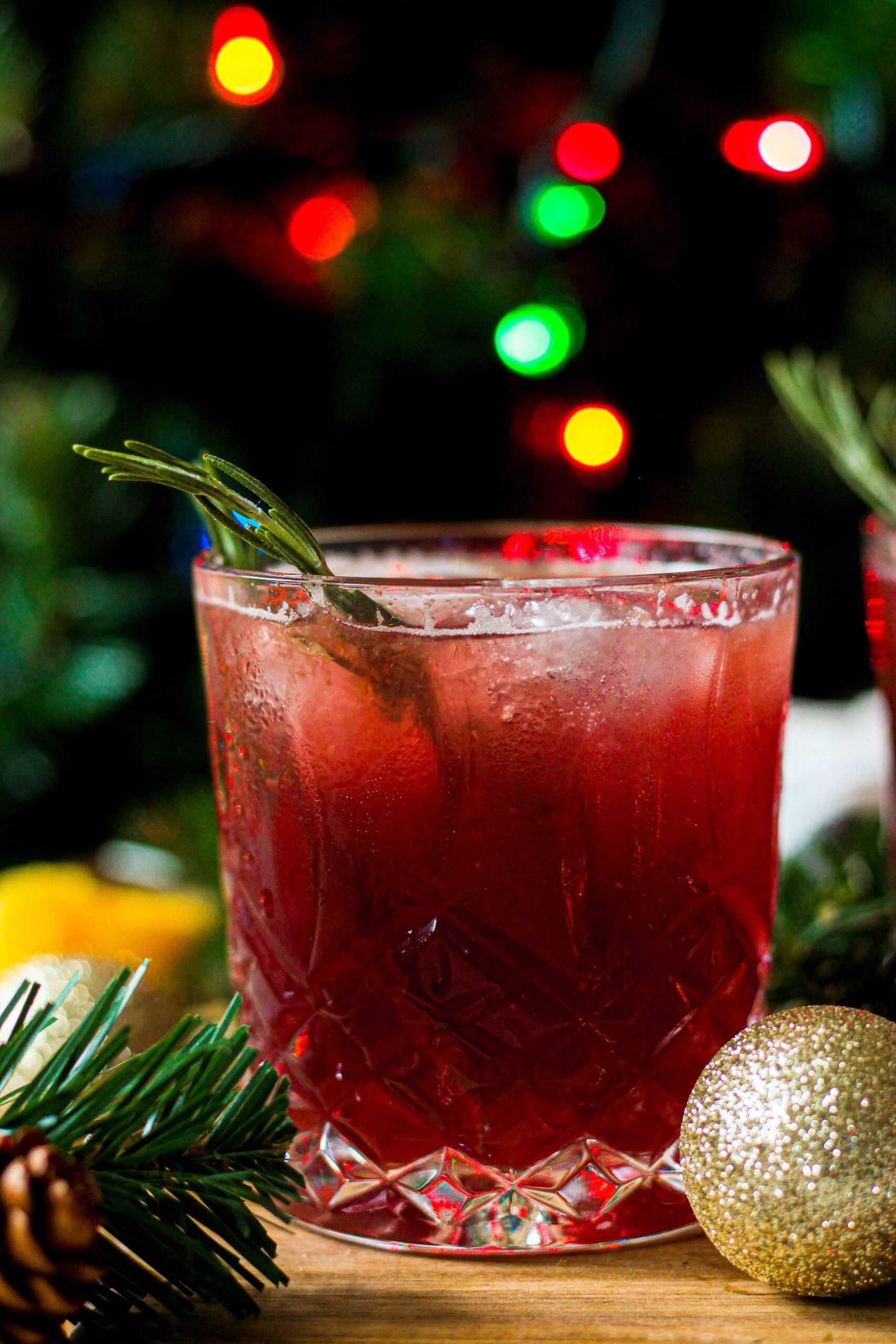 A glass of red cocktail garnished with a sprig of rosemary sits on a wooden surface, surrounded by pine branches and a gold ornament, with colorful lights blurred in the background.
