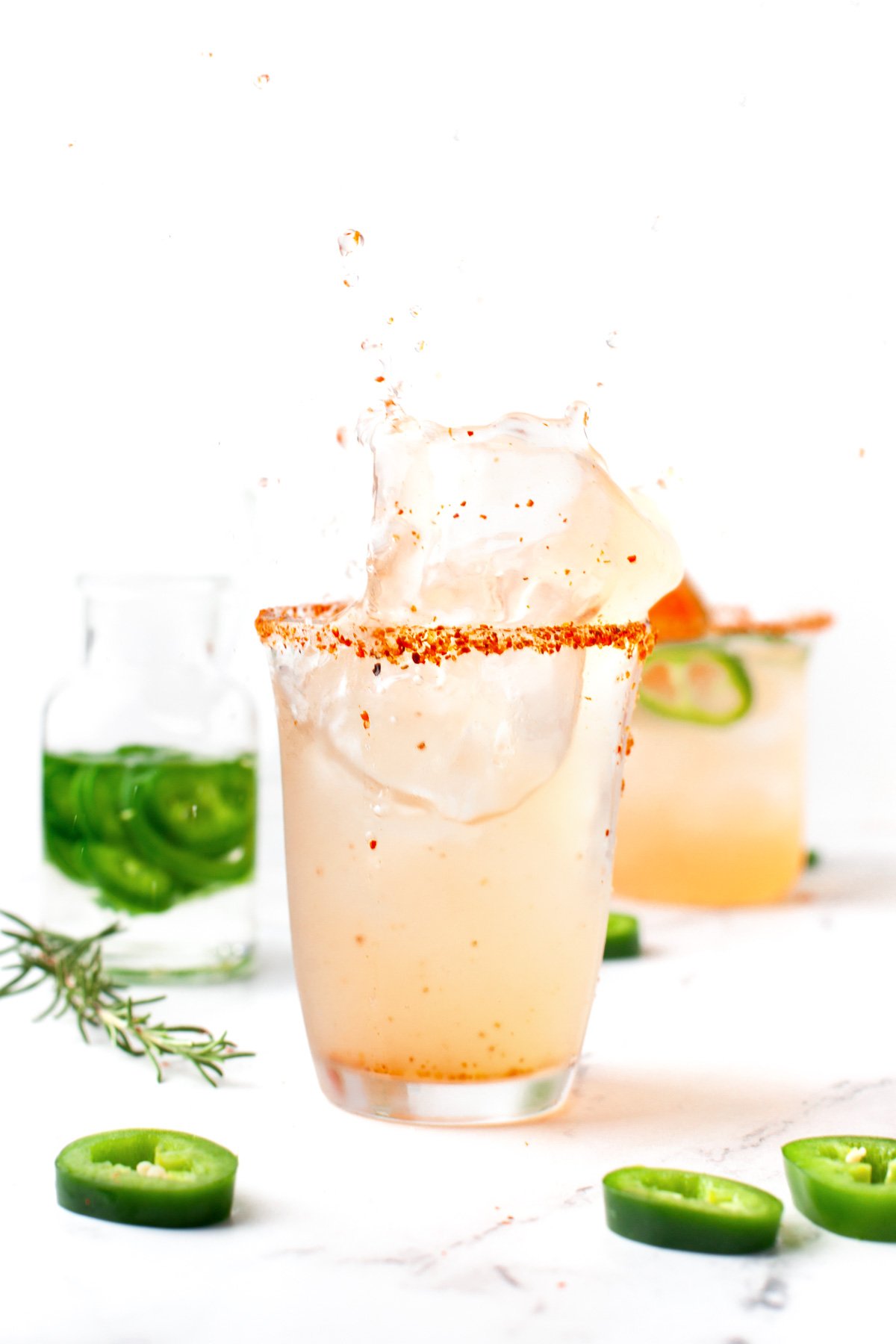 A glass of light-colored cocktail with a chili powder rim and ice splash, surrounded by sliced jalapeños and a jar of jalapeño slices on a white surface.