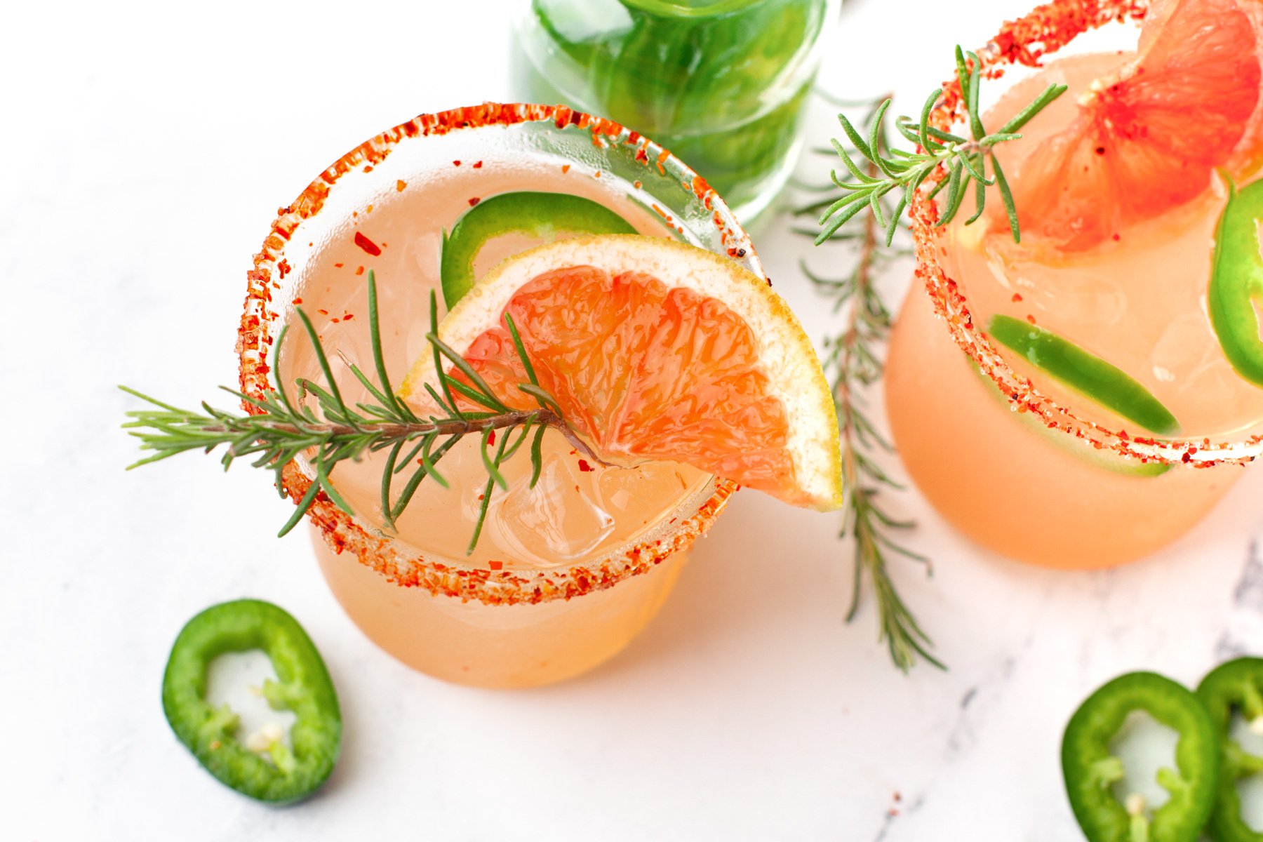 Two grapefruit cocktails with chili powder rims, garnished with rosemary, jalapeño slices, and grapefruit wedges, on a white surface with ice and extra jalapeño slices nearby.