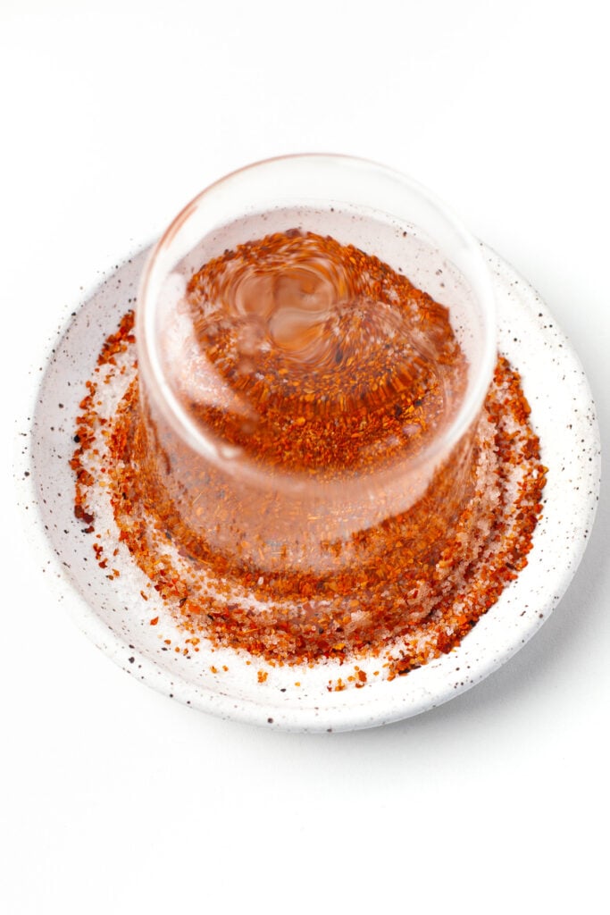 A glass is being dipped upside down into chili powder on a white plate to coat its rim.