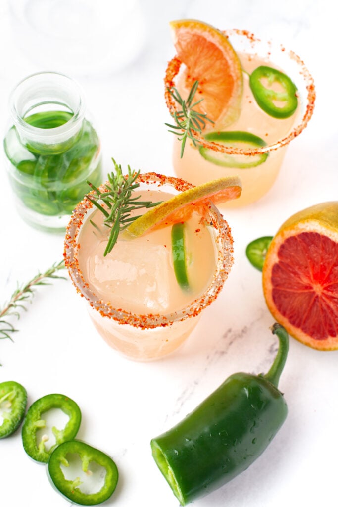 Two glasses of a light pink cocktail garnished with rosemary, grapefruit slice, and jalapeño, with a chili powder rim. Sliced jalapeño and grapefruit are nearby.
