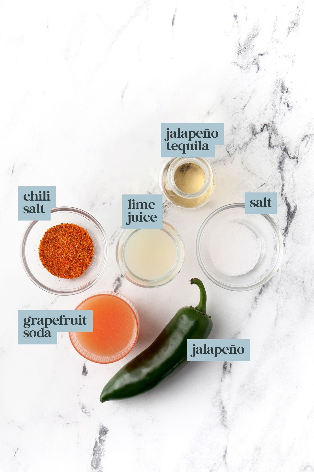 Five labeled ingredients on a white surface: grapefruit soda, chili salt, lime juice, jalapeño tequila, salt, and a whole jalapeño.