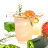 A glass of light-colored spicy paloma cocktail garnished with a jalapeño slice, grapefruit slice, rosemary sprig, and a chili powder rim, with fresh jalapeños and rosemary nearby.
