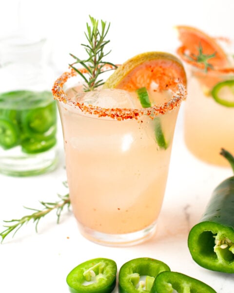 A spicy paloma cocktail garnished with a rosemary sprig, grapefruit slice, and jalapeño ring sits in a glass with a chili powder rim; sliced jalapeños and another drink appear in the background.