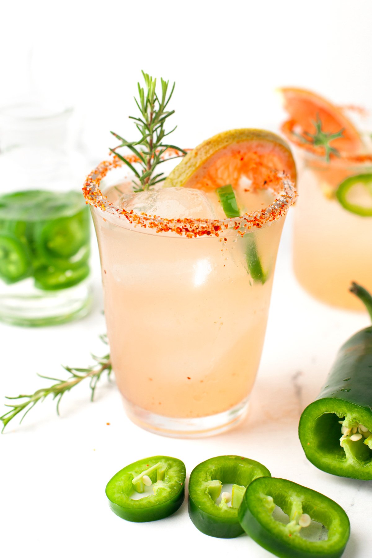 A spicy paloma cocktail garnished with a rosemary sprig, grapefruit slice, and jalapeño ring sits in a glass with a chili powder rim; sliced jalapeños and another drink appear in the background.