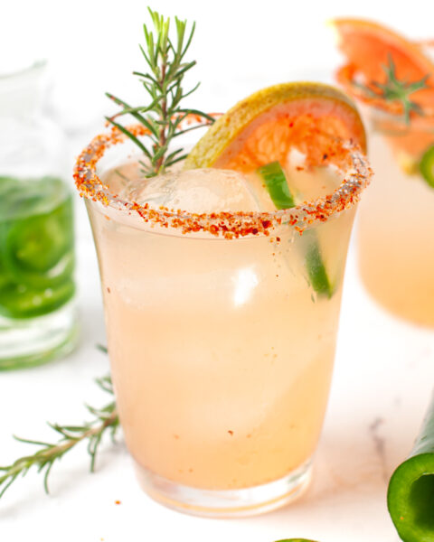 A glass of light pink spicy Paloma cocktail with ice, a chili powder rim, garnished with a grapefruit slice, rosemary sprig, and jalapeño slices, sits on a white surface.