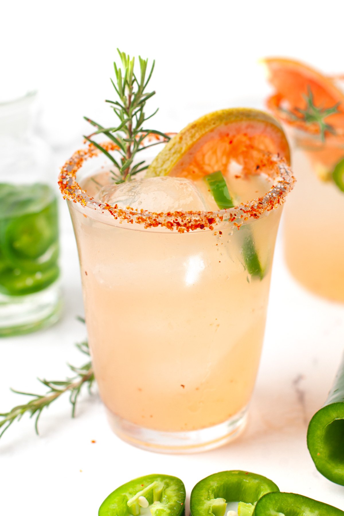 A glass of light pink spicy Paloma cocktail with ice, a chili powder rim, garnished with a grapefruit slice, rosemary sprig, and jalapeño slices, sits on a white surface.