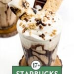 A glass of Starbucks-style iced drink topped with whipped cream, chocolate drizzle, a chocolate piece, graham cracker, toasted marshmallow, and crushed toppings. Text reads "17+ Starbucks Drinks.