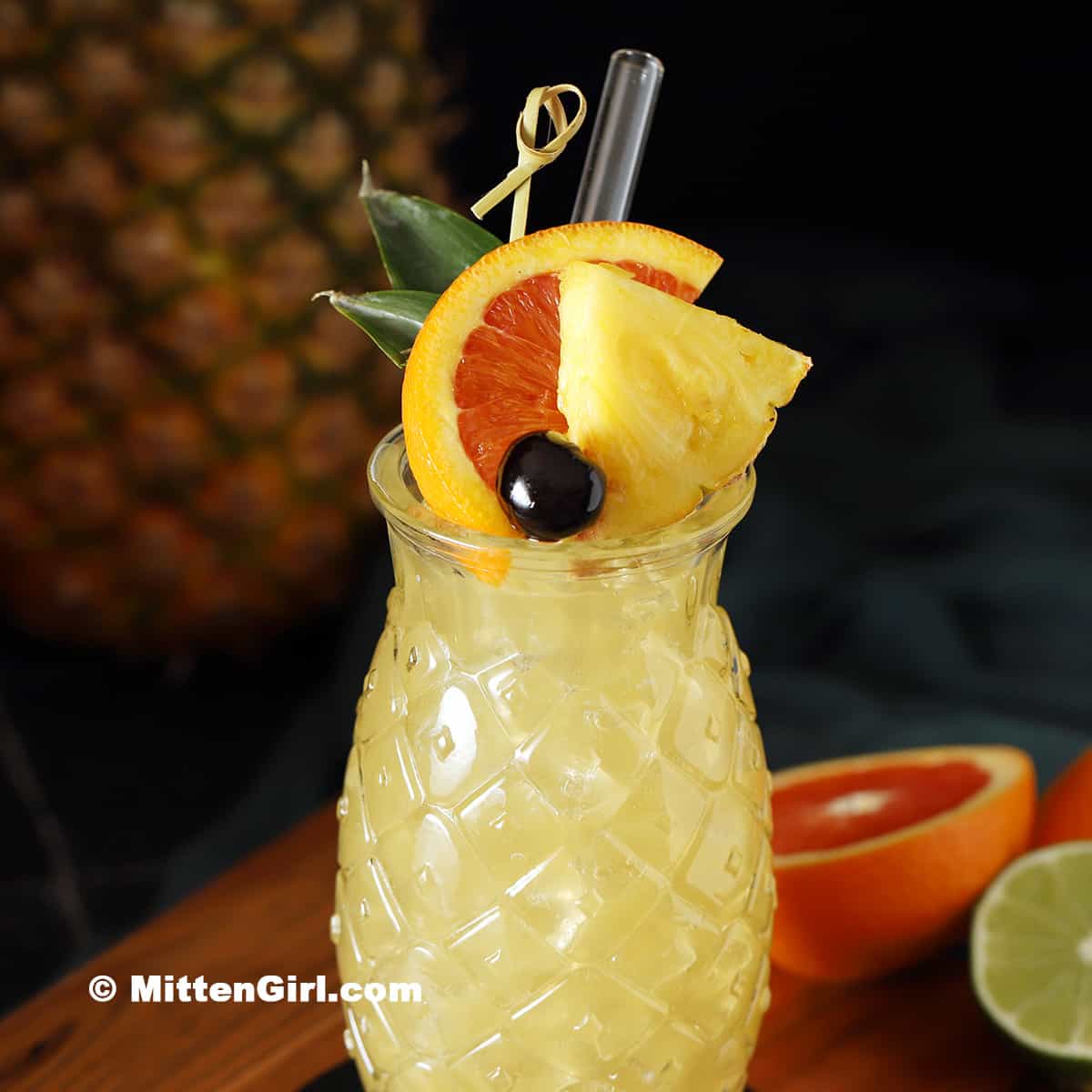 A tropical cocktail in a pineapple-patterned glass, garnished with a pineapple wedge, orange slice, cherry, and straw. Pineapple and citrus fruits are in the background.