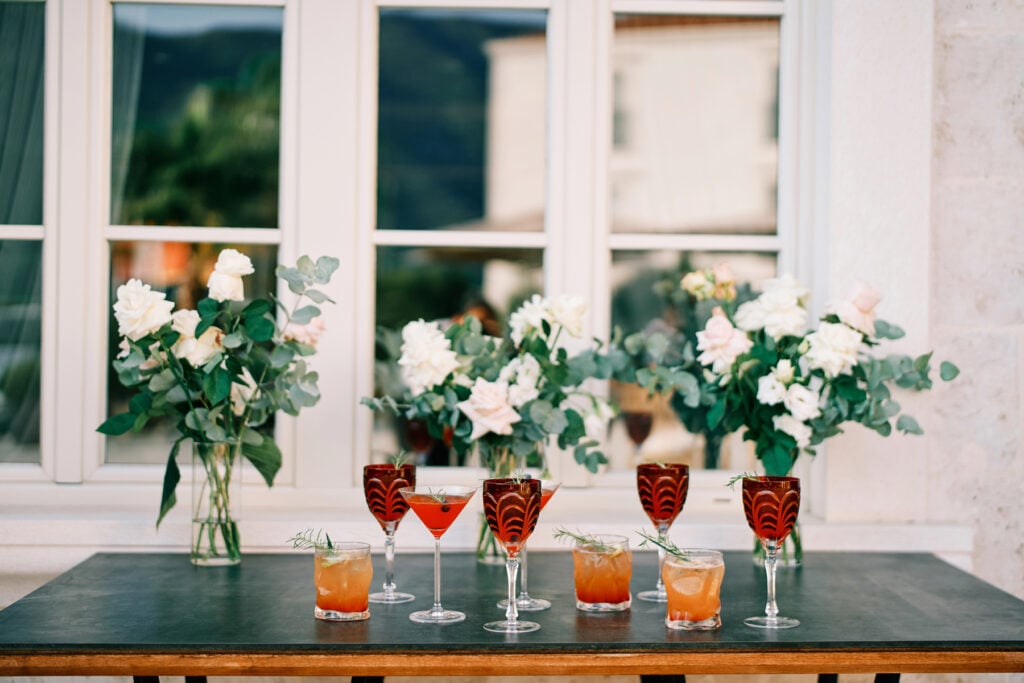 A table with six signature cocktails for weddings in decorative glasses, garnished with herbs, is set in front of a window with three vases of white and pink flowers.