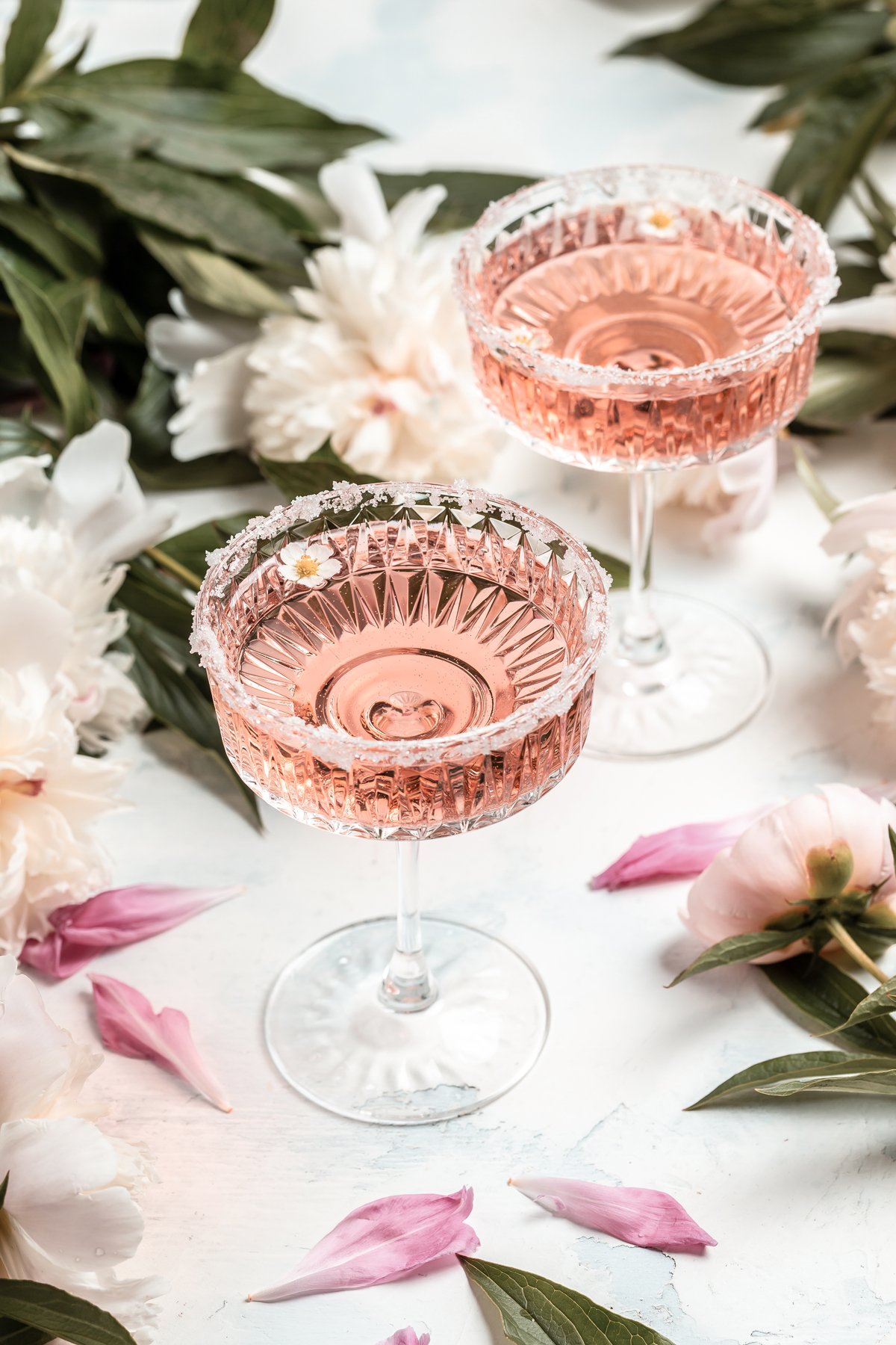 Two elegant pink cocktails in vintage glasses with sugared rims, surrounded by white flowers, greenery, and pink petals—perfect as signature cocktails for weddings.