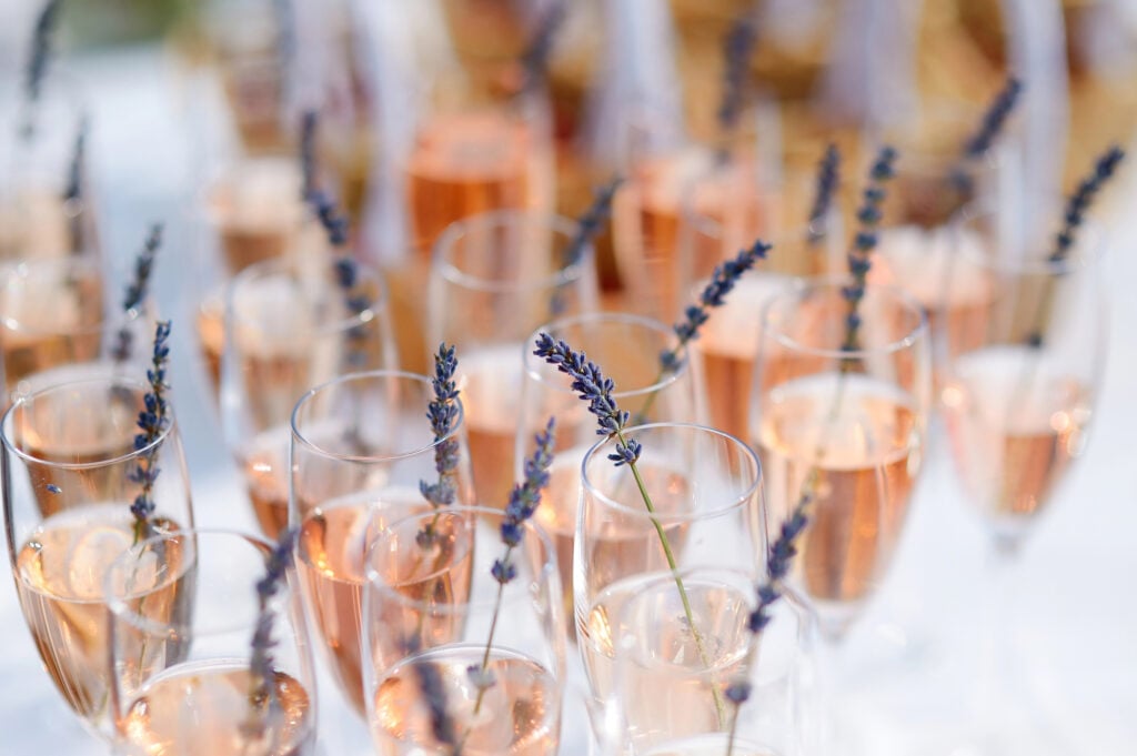 Rows of champagne flutes filled with rosé sparkling wine, each glass garnished with a sprig of lavender, create elegant signature cocktails for weddings, beautifully arranged on a white surface.