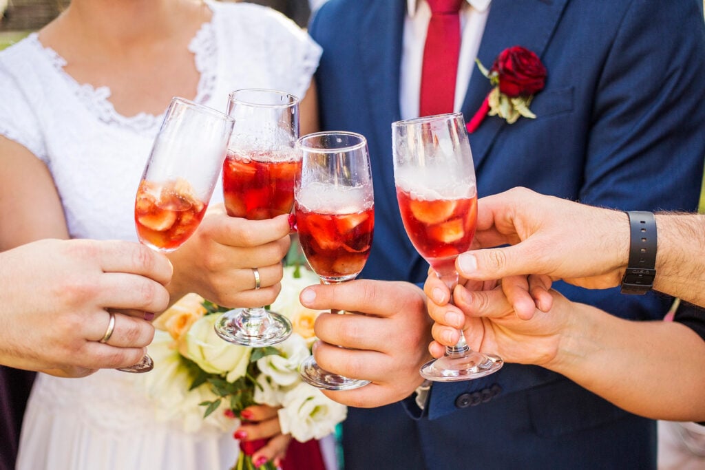 Several people, some in wedding attire, clink glasses filled with signature cocktails for weddings—a vibrant red iced beverage—in a joyful toast; one person holds a bouquet of white roses.