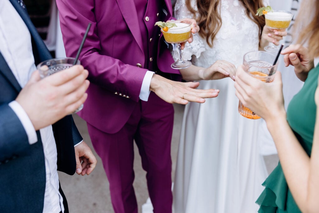 Four people in formal attire stand together holding signature cocktails for weddings; one person in a purple suit gestures with their hand during conversation.