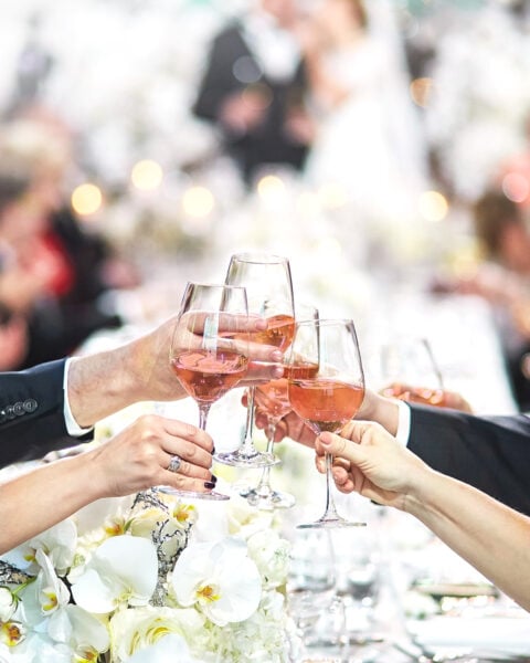 Five people clink glasses of rosé wine over a table decorated with white flowers, celebrating with Signature Cocktails for Weddings as a blurred wedding scene unfolds in the background.