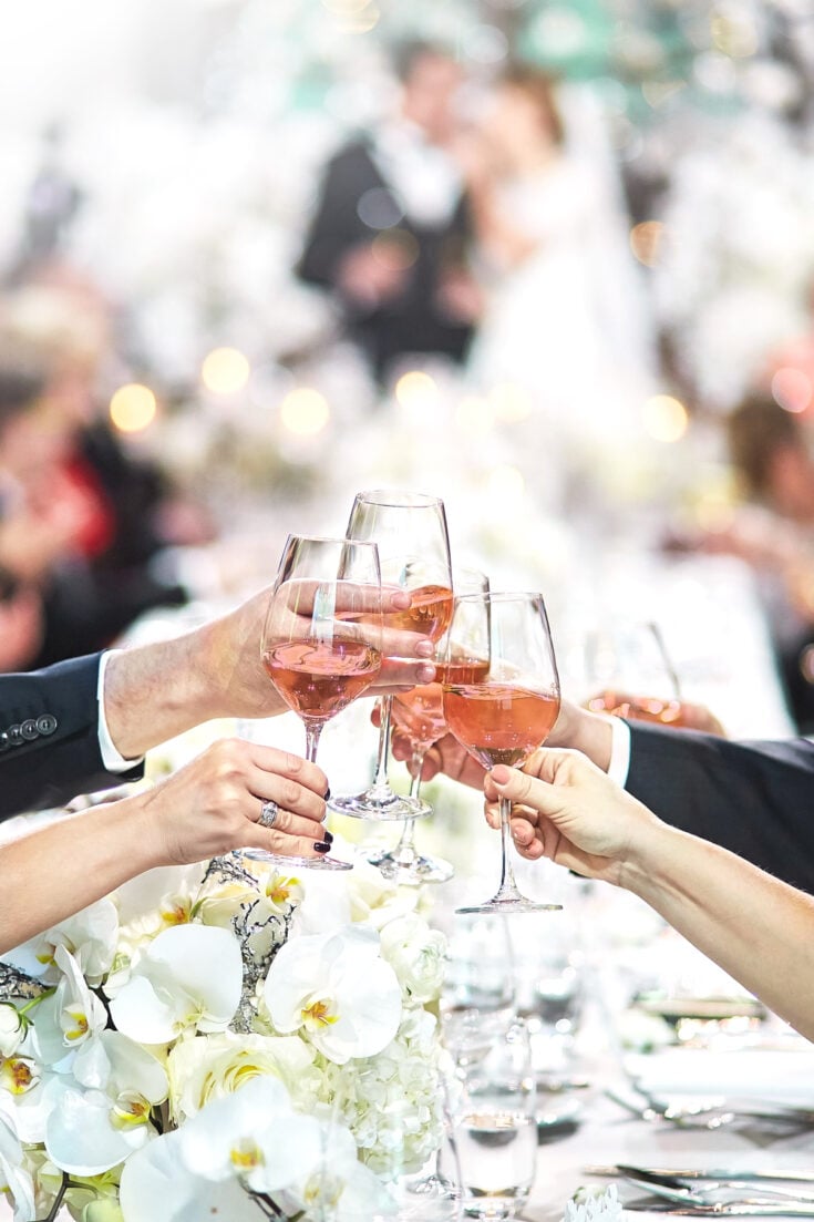 Five people clink glasses of rosé wine over a table decorated with white flowers, celebrating with Signature Cocktails for Weddings as a blurred wedding scene unfolds in the background.