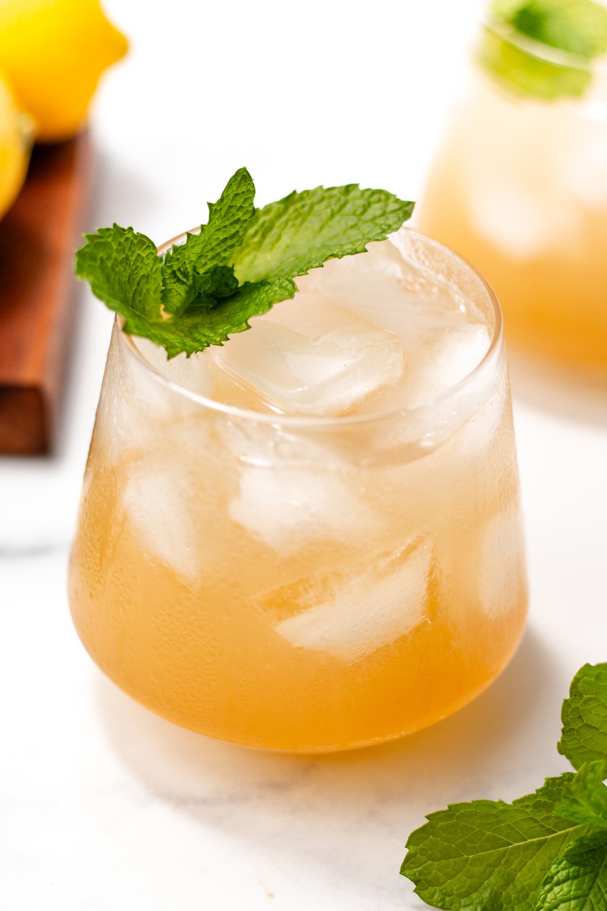A glass of iced light brown cocktail garnished with a mint sprig, with lemon and mint leaves in the background on a white surface.