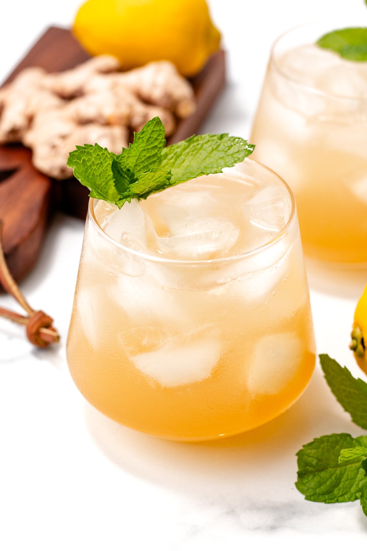 A glass of iced light brown beverage garnished with a mint sprig, with ginger, lemon, and another glass in the background.