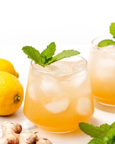 Two glasses of iced ginger lemonade garnished with mint, with fresh lemons, ginger, and mint leaves placed nearby on a white surface.