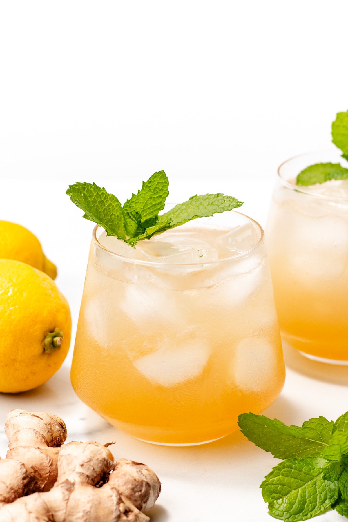 Two glasses of iced ginger lemonade garnished with mint, with fresh lemons, ginger, and mint leaves placed nearby on a white surface.