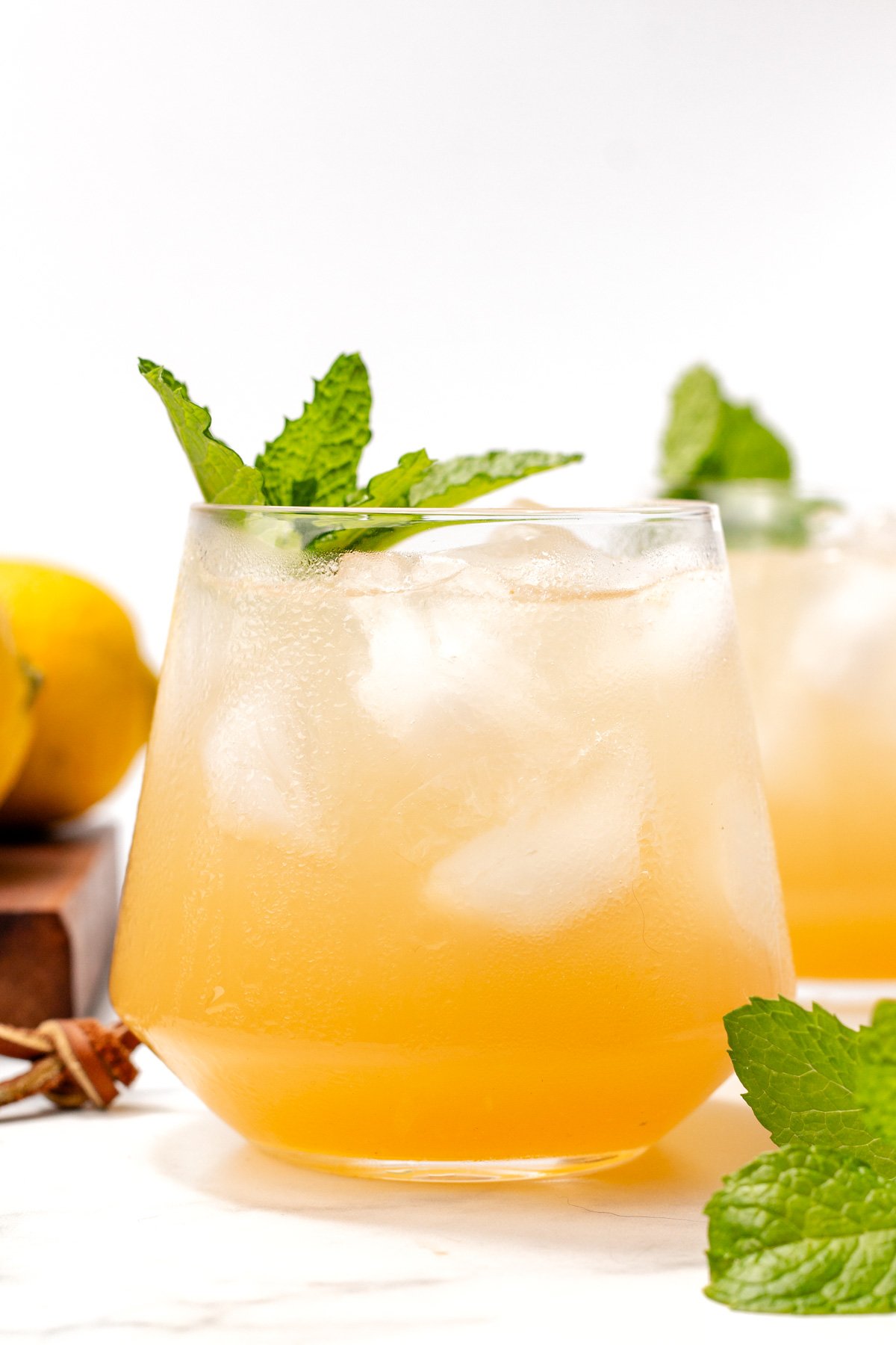 A glass of iced yellow-orange beverage garnished with fresh mint leaves, with another similar drink and a lemon in the background.