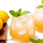 A glass of iced lemonade garnished with a fresh mint sprig, with lemons and another glass in the background on a white surface.