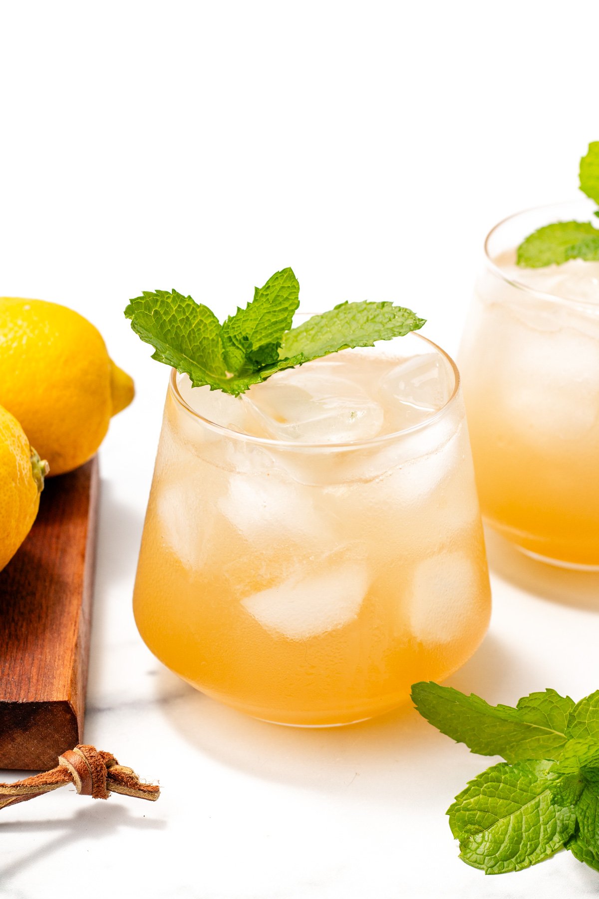 A glass of iced lemonade garnished with a fresh mint sprig, with lemons and another glass in the background on a white surface.