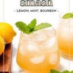A glass of whiskey smash cocktail with ice, garnished with a mint sprig, sits beside lemons and another glass on a white surface. Text reads: "whiskey smash – lemon, mint, bourbon.