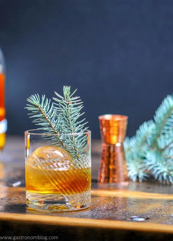 A cocktail in a glass with a large round ice cube and a pine sprig garnish, inspired by old-fashioned-cocktails, sits on a wooden surface with a copper jigger and another pine sprig in the background.