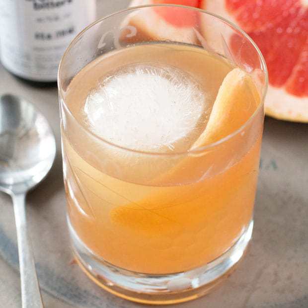A glass of light orange cocktail with a large round ice cube and a grapefruit twist, placed next to a spoon and a halved grapefruit.