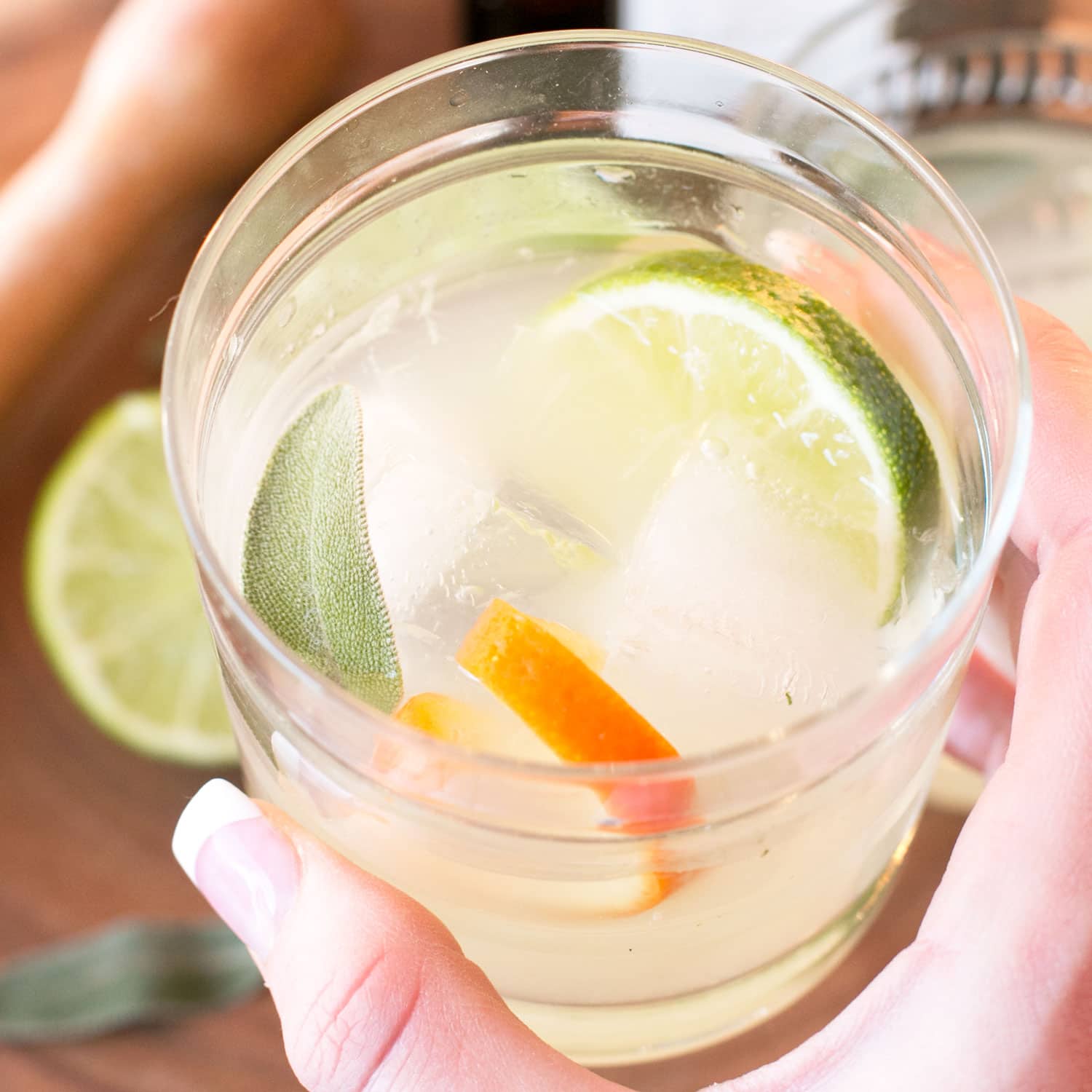 A hand holds a glass with a clear cocktail containing ice cubes, lime and orange slices, and a sage leaf. Another lime slice is visible on the table in the background.