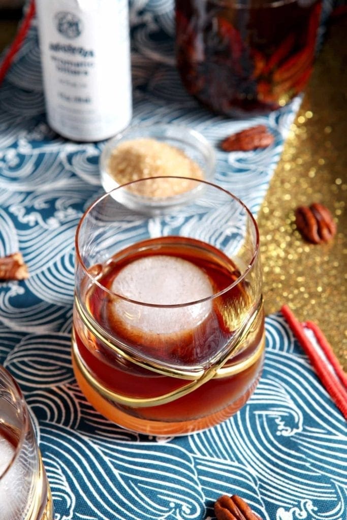 A glass of brown old-fashioned-cocktail with a large ice cube sits on a blue patterned cloth, surrounded by pecans, a bowl of brown sugar, and a canned beverage in the background.
